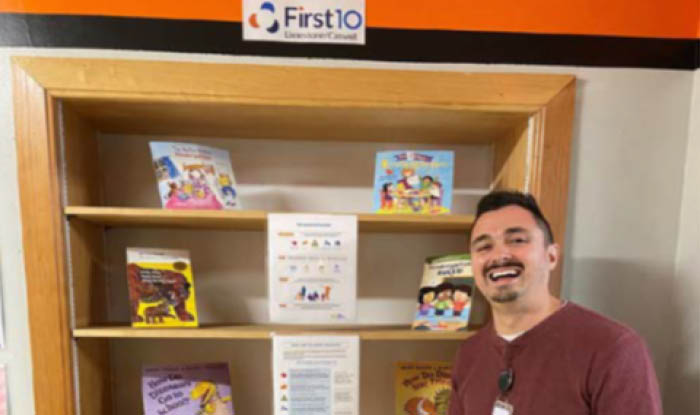 Bookshelf of selected books for young children and a First 10 sign, with a First 10 Sanford team member standing in front of the bookshelf.