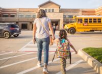 A mother holding the hand of a child as they walk towards a school building.