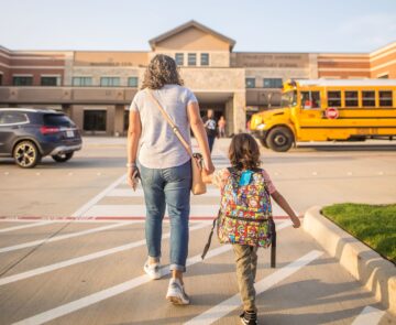 A mother holding the hand of a child as they walk towards a school building.