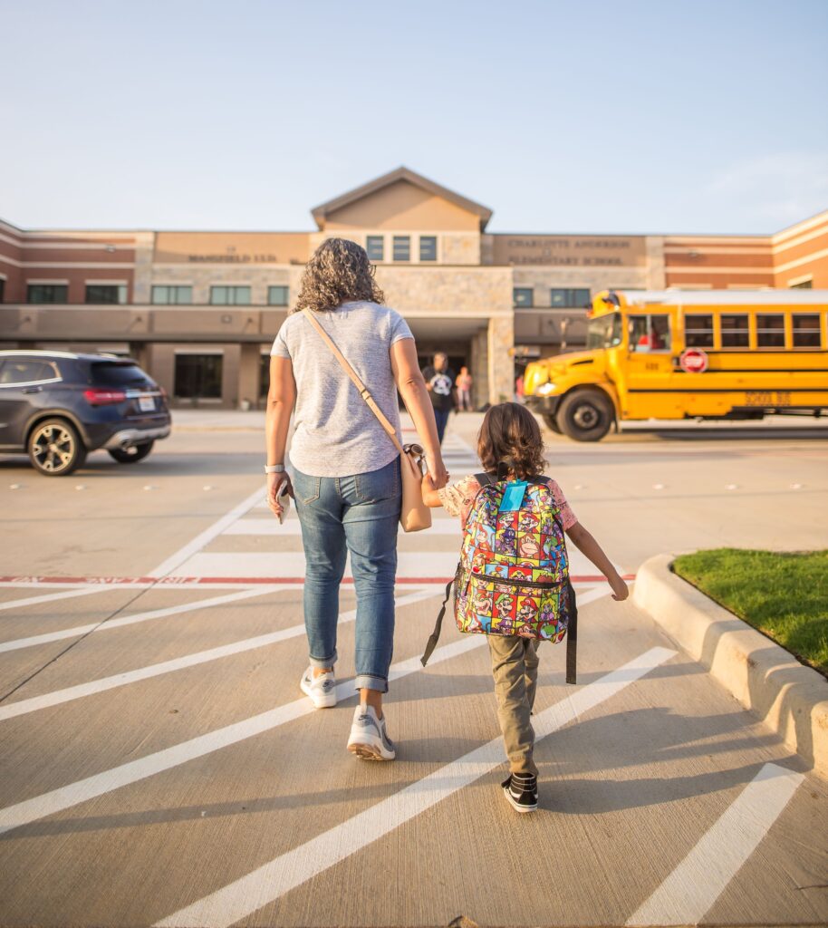 A mother holding the hand of a child as they walk towards a school building.